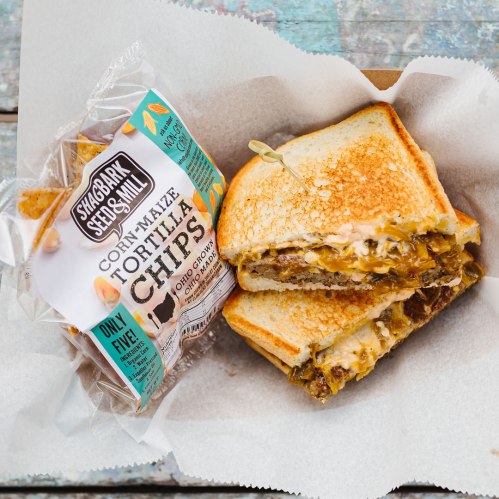 A toasted sandwich filled with melted cheese and toppings, served with a side of tortilla chips in a paper container.