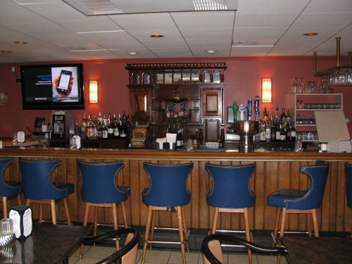 A bar area with blue chairs, a wooden counter, shelves of liquor, and a TV on the wall.