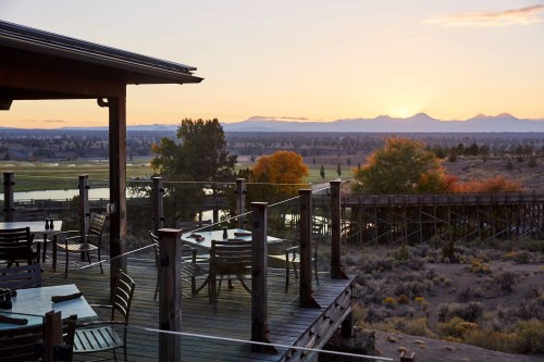A wooden deck with tables overlooks a scenic landscape at sunset, featuring mountains and autumn foliage.