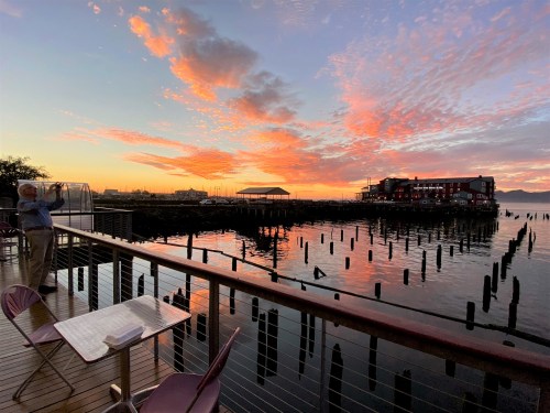 A serene sunset over water, with vibrant clouds and a silhouette of a man on a deck, reflecting on the calm scene.