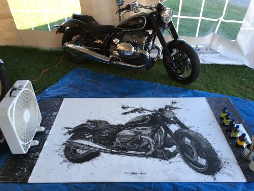 A black and white motorcycle painting on a blue tarp, with a fan and paint bottles nearby, and a motorcycle in the background.