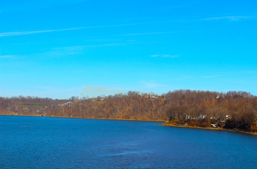 A serene lake surrounded by trees under a clear blue sky, with distant houses on the hillside.