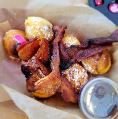 A basket of golden-brown doughnuts topped with crispy bacon and powdered sugar, served with a side of dipping sauce.