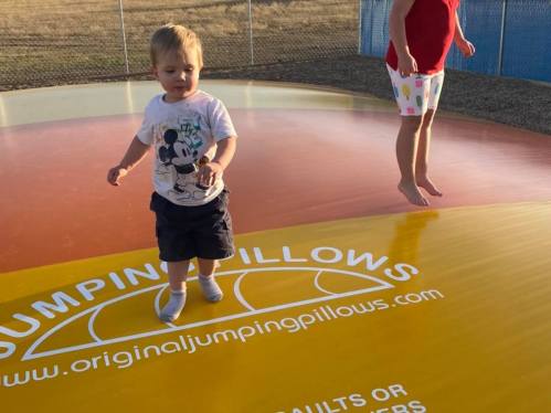 A toddler in a Mickey Mouse shirt plays on a colorful jumping pillow while an older child jumps nearby.