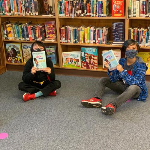 Two children sitting on a carpet in a library, each holding a book and smiling, surrounded by shelves of books.