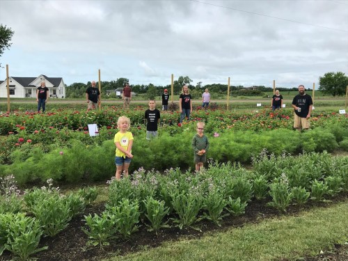 A group of children and adults stand in a vibrant flower garden, surrounded by colorful blooms and greenery.
