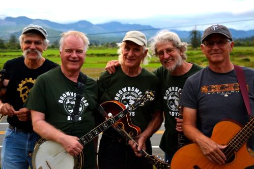 Five musicians in matching shirts pose outdoors with instruments, set against a scenic landscape of mountains and fields.