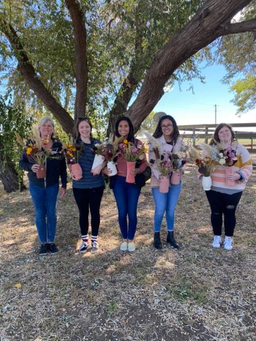 Five women stand together outdoors, each holding a bouquet of dried flowers in pots, with trees in the background.