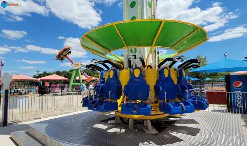 A colorful amusement park ride with blue seats and a green canopy, set against a bright blue sky and nearby attractions.