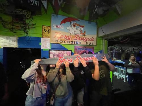 A group of four people playfully hiding their faces in front of the Tipsy Seagull Dockside Pub sign.