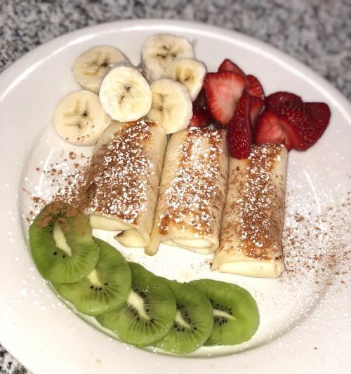 Three crepes dusted with powdered sugar, served with banana slices, strawberries, and kiwi on a white plate.