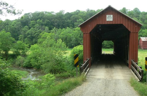 A red covered bridge spans a small river, surrounded by lush green trees and fields.