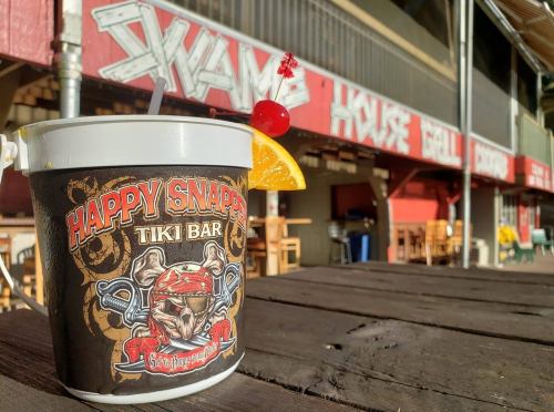 A colorful drink in a bucket with "Happy Snapper Tiki Bar" logo, set against a rustic bar backdrop.