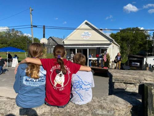 Three children sit on a stone wall, watching a performance at a country store on a sunny day.