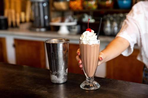 A hand holds a tall glass of chocolate milkshake topped with whipped cream and a cherry, with a metal shaker nearby.