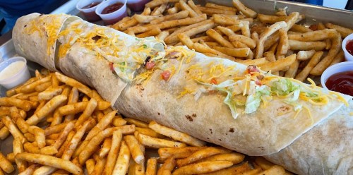 A large burrito topped with cheese and lettuce, served with a side of golden French fries and dipping sauces.