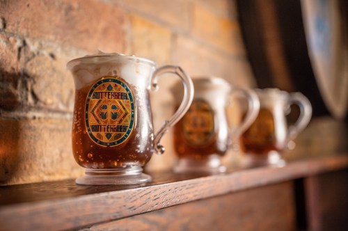 Three frosty mugs of butterbeer sit on a wooden shelf against a brick wall, each adorned with a colorful label.