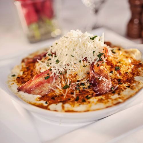 A plate of creamy pasta topped with grated cheese, herbs, and crispy bacon, served elegantly on a white dish.