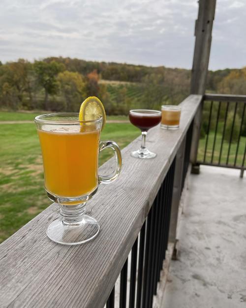 Three cocktails on a wooden railing, with a scenic view of green fields and cloudy skies in the background.