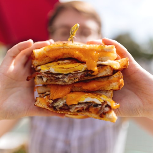A person holds a towering sandwich filled with layers of cheese, eggs, and bacon against a sunny background.