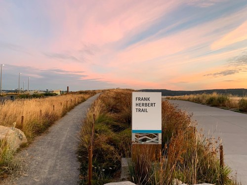 A sign for the Frank Herbert Trail beside a path, with a colorful sunset sky and grassy surroundings.