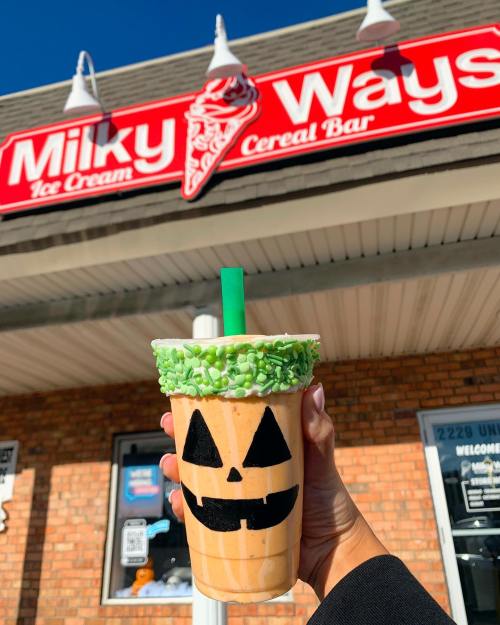 A hand holds a Halloween-themed milkshake with a jack-o'-lantern face and green sprinkles, in front of a shop sign.