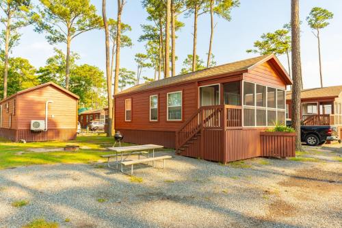 A cozy cabin with a porch surrounded by tall trees, set in a gravel area with a picnic table and nearby vehicles.