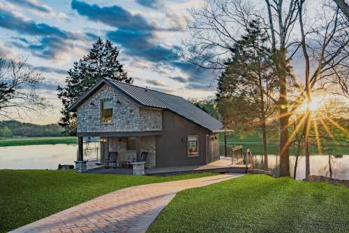 A modern house by a serene lake, surrounded by trees, with a pathway leading to it and a sunset in the background.