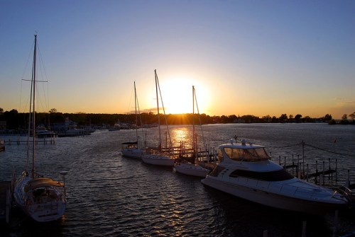 Sunset over a calm harbor with several boats docked, casting reflections on the water.