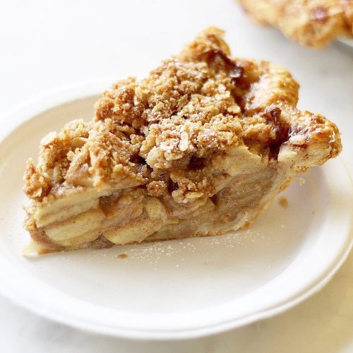 A slice of apple pie topped with a crumbly oat topping, served on a white plate.
