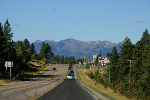 A scenic road leads towards mountains, with cars driving and trees lining the sides under a clear blue sky.