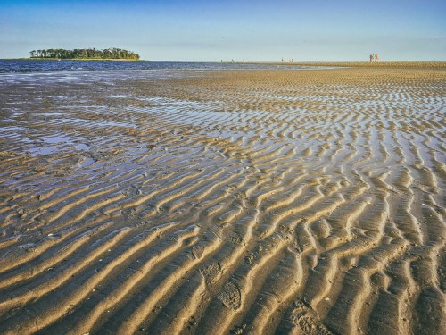 A sandy beach with rippled patterns, leading to a calm sea and a distant island under a clear blue sky.
