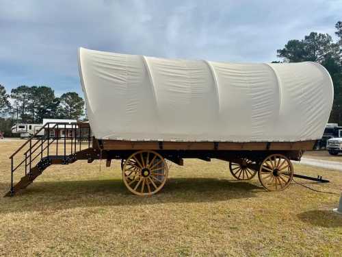 A covered wagon with a white canvas top, resting on large wooden wheels, set on grassy ground.