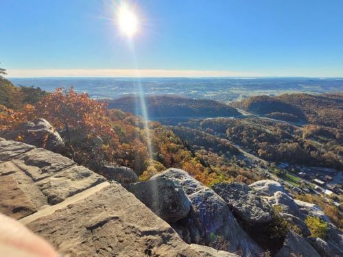 A scenic view from a rocky overlook, showcasing rolling hills and a bright sun in a clear blue sky.