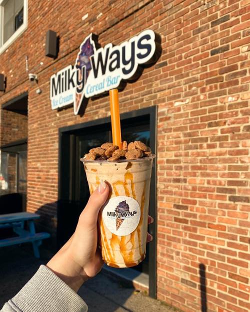 A hand holding a milkshake topped with cereal and caramel, in front of a brick wall with a "Milky Ways" sign.