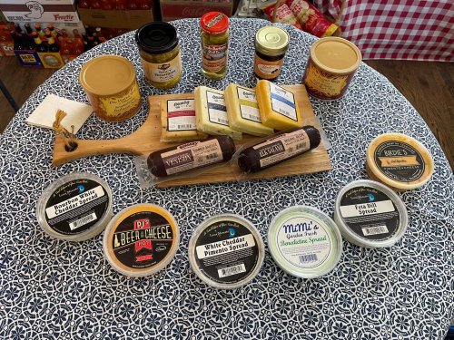 A wooden board with various cheeses and spreads, surrounded by jars and containers of condiments on a patterned tablecloth.