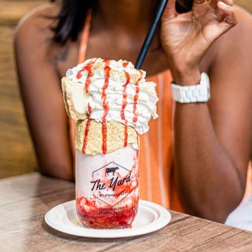A tall milkshake topped with whipped cream, cake, and strawberry sauce, served in a glass with a straw.