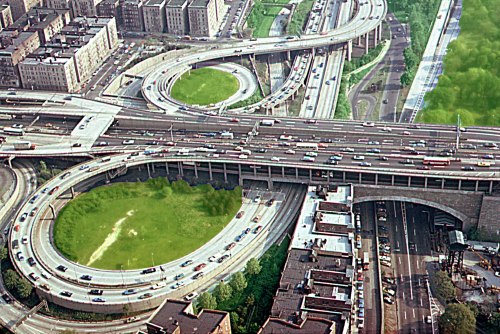 Aerial view of a complex highway interchange with multiple lanes and green spaces amidst urban buildings.