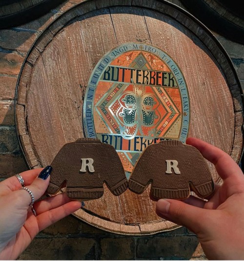 Two hands hold brown coasters shaped like sweaters, featuring the letter "R," in front of a decorative Butterbeer barrel.