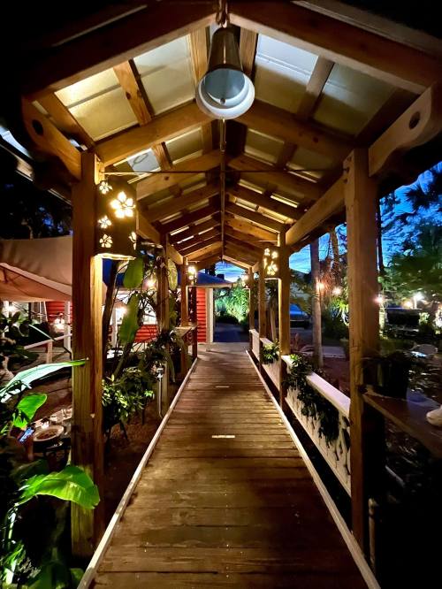 A wooden walkway illuminated by soft lights, leading through a lush garden to a cozy outdoor dining area.