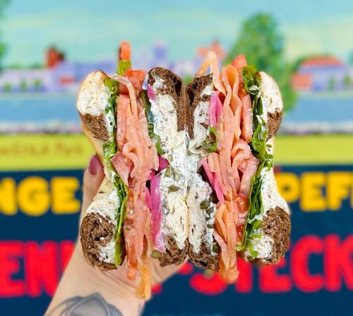 A hand holding a cross-section of a colorful sandwich with layers of vegetables and cream cheese on dark bread.