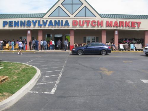 A busy Pennsylvania Dutch Market with a long line of people outside and a parked car in the foreground.