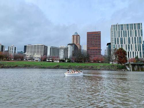A small boat with people is on a river, with a city skyline and green park in the background under a cloudy sky.