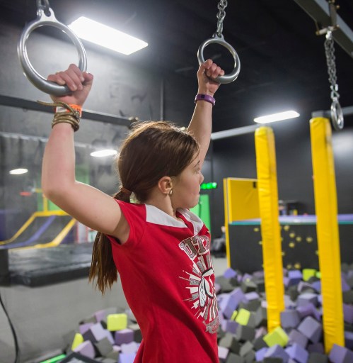 A girl hangs from gymnastic rings in a trampoline park, surrounded by colorful foam blocks.