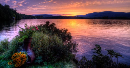 A serene lakeside scene at sunset, with colorful flowers and mountains in the background reflecting on the water.