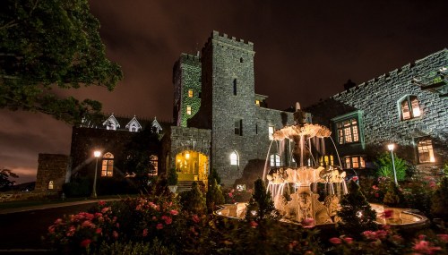 A stone castle at night, featuring a fountain and illuminated gardens, surrounded by trees and a cloudy sky.