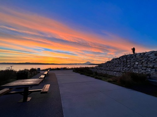 A scenic sunset with vibrant clouds over a waterway, featuring a silhouette of a person on a rocky hill.