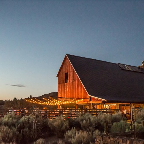 A rustic barn illuminated by string lights at dusk, surrounded by sagebrush and a clear sky.