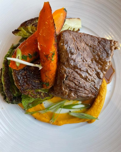 A plated dish featuring a piece of beef, roasted carrots, and greens on a bed of orange puree.