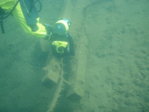 A diver in yellow gear examines a submerged object on the ocean floor, with a robotic device nearby.